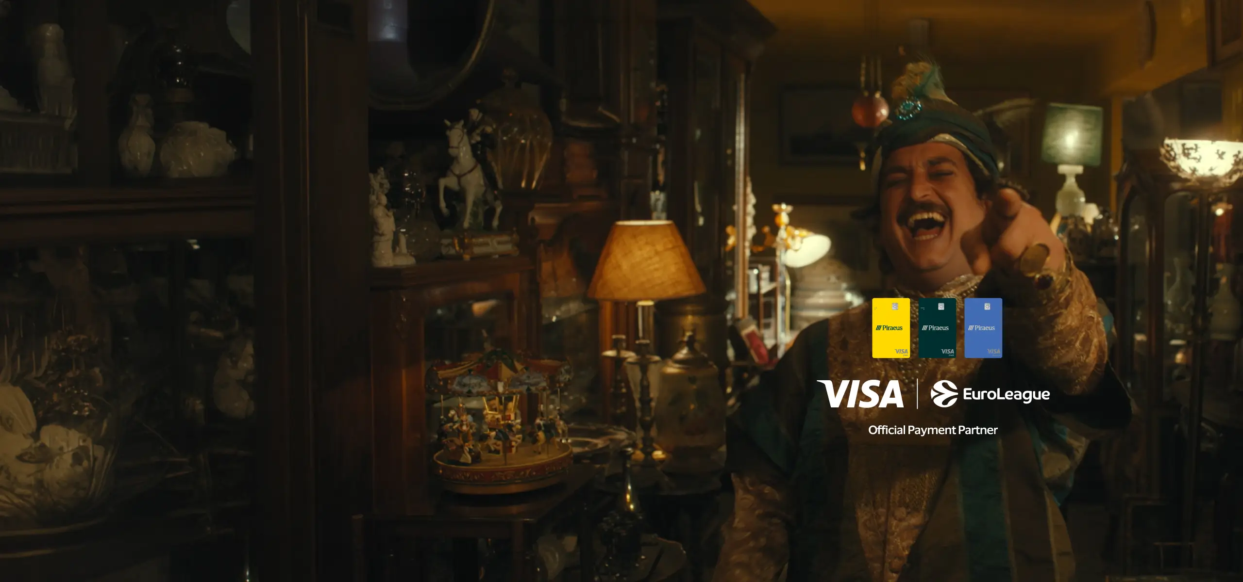 Άνδρας ντυμένος με παραδοσιακή στολή γελά και δείχνει προς την κάμερα σε ζεστό φωτισμένο εσωτερικό χώρο, με λογότυπα Visa και EuroLeague