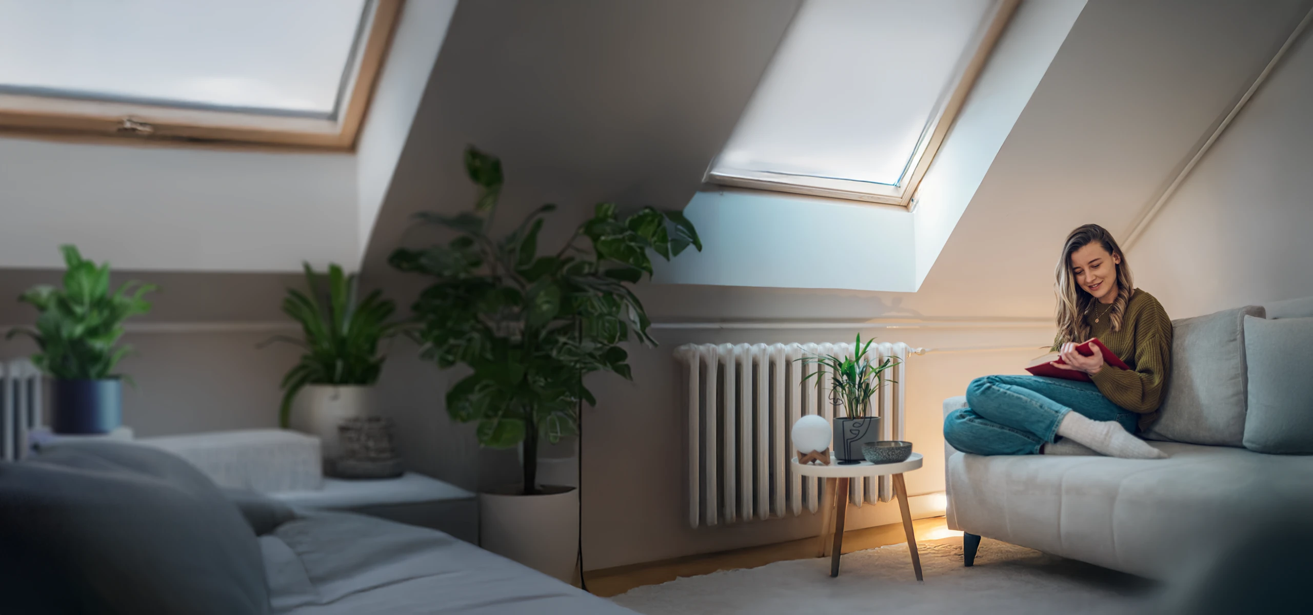 Woman sitting comfortably on a sofa in a bright attic room, reading a book near a skylight and indoor plants.